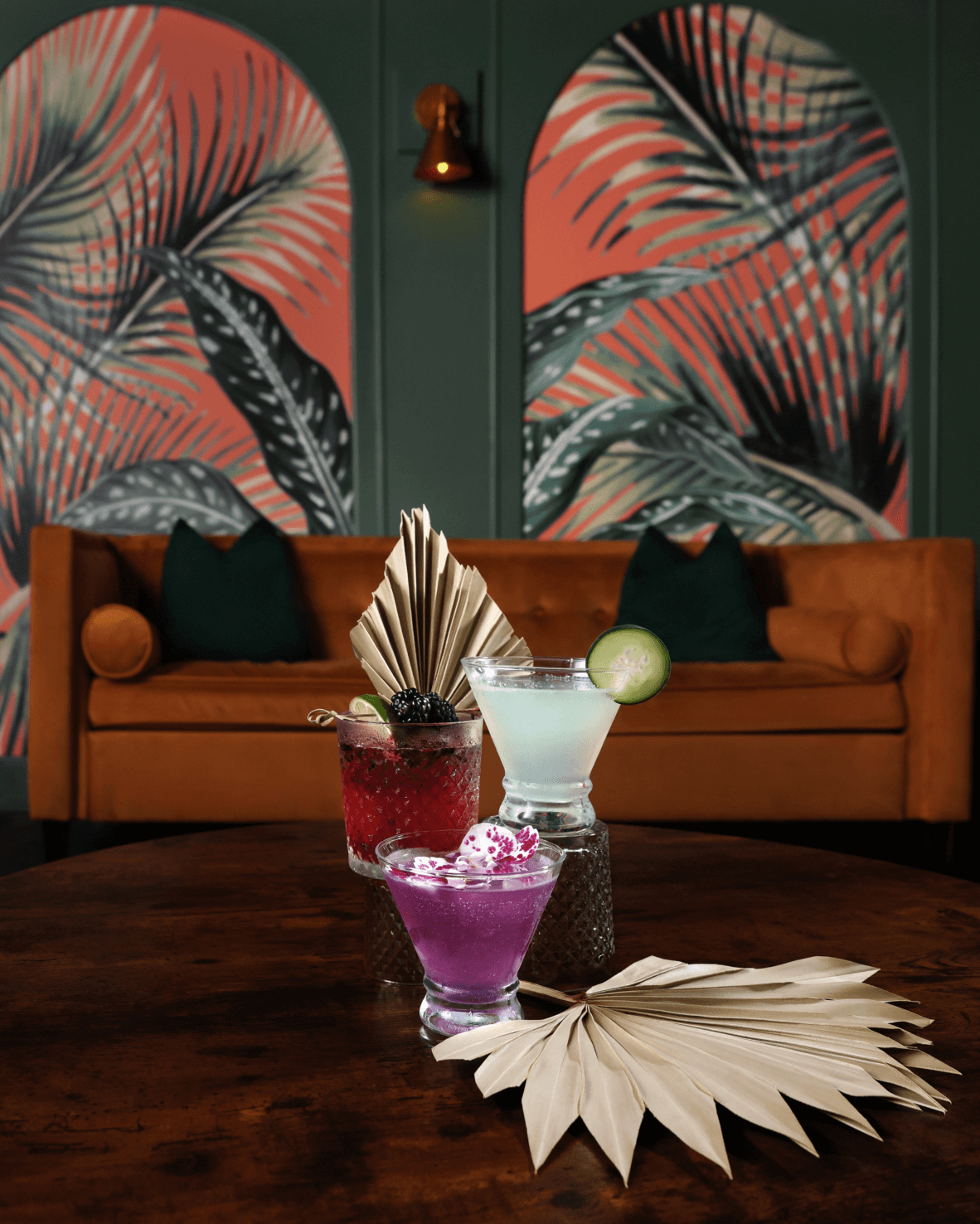 Palm Social Club cocktails on a velvet sofa with palm-leaf wallpaper